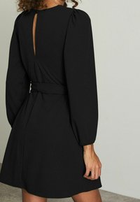 Svart klänning med puffärmar, midjebälte och en nyckelhålsdetalj på baksidan. Mjuk tyg och slät textur, knälång design.