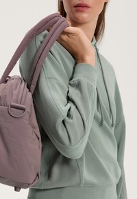 Puha, zöld kapucnis pulóver bordázott textúrával és húzózsinórral. Szürke-lila válltáska lekerekített formával és párnázott pántokkal, amelyet a viselője tart.