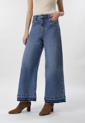 Frau trägt hochtaillierte, weit geschnittene blaue Jeans mit ausgefransten Säumen, kombiniert mit braunen Stiefeletten mit Absatz und einem weißen ärmellosen Oberteil.