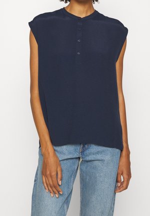 Personne portant un haut sans manches bleu marine boutonné et un jean bleu clair, debout devant un fond blanc uni.