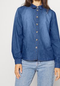 Jeansskjorta i mörkblått med krage, långa ärmar med figurnära manschetter och bruna knappar framtill. Enkel, rak skärning.