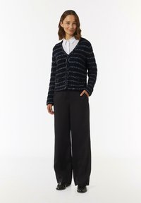 Strukturierter marineblauer Cardigan mit hellblauen Streifen, V-Ausschnitt und Knopfverschluss. Kombiniert mit weiten schwarzen Hosen und glänzenden schwarzen Schuhen.