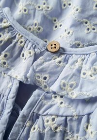 Lyseblå kjole i stof med hvid blomstret broderi, med flæsekant og en træknapdetalje ved halsudskæringen.