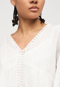 Κοντινό πλάνο ενός ατόμου που φοράει μια λευκή υφασμάτινη μπλούζα με δαντέλα κατά μήκος του V-λαιμού και της μπροστινής ραφής.