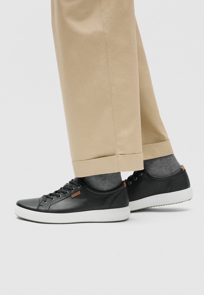 Zapatillas bajas de piel negra con suelas blancas, usadas con calcetines grises y pantalones beige con puños sobre un fondo gris claro.