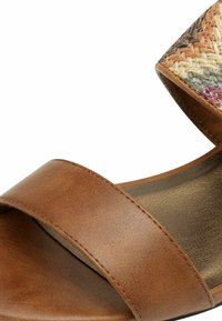 Sandale en cuir marron avec de larges sangles croisées et un motif tissé texturé sur le talon. La surface intérieure lisse réfléchit la lumière.