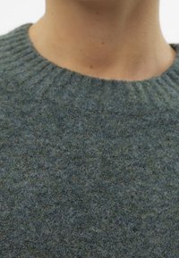 Läheltä otettu kuva kaulasta ja hartioista, joissa on päällä teksturoitu, ribattu pyöreäkauluksinen villapaita, valmistettu kirjavasta tumman vihreästä ja sinisestä kankaasta.