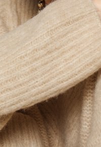 Beżowy sweter dziany z detalami, który charakteryzuje się prążkowaną strukturą, miękkim wełnianym materiałem i akcentem w postaci złożonego rękawa.