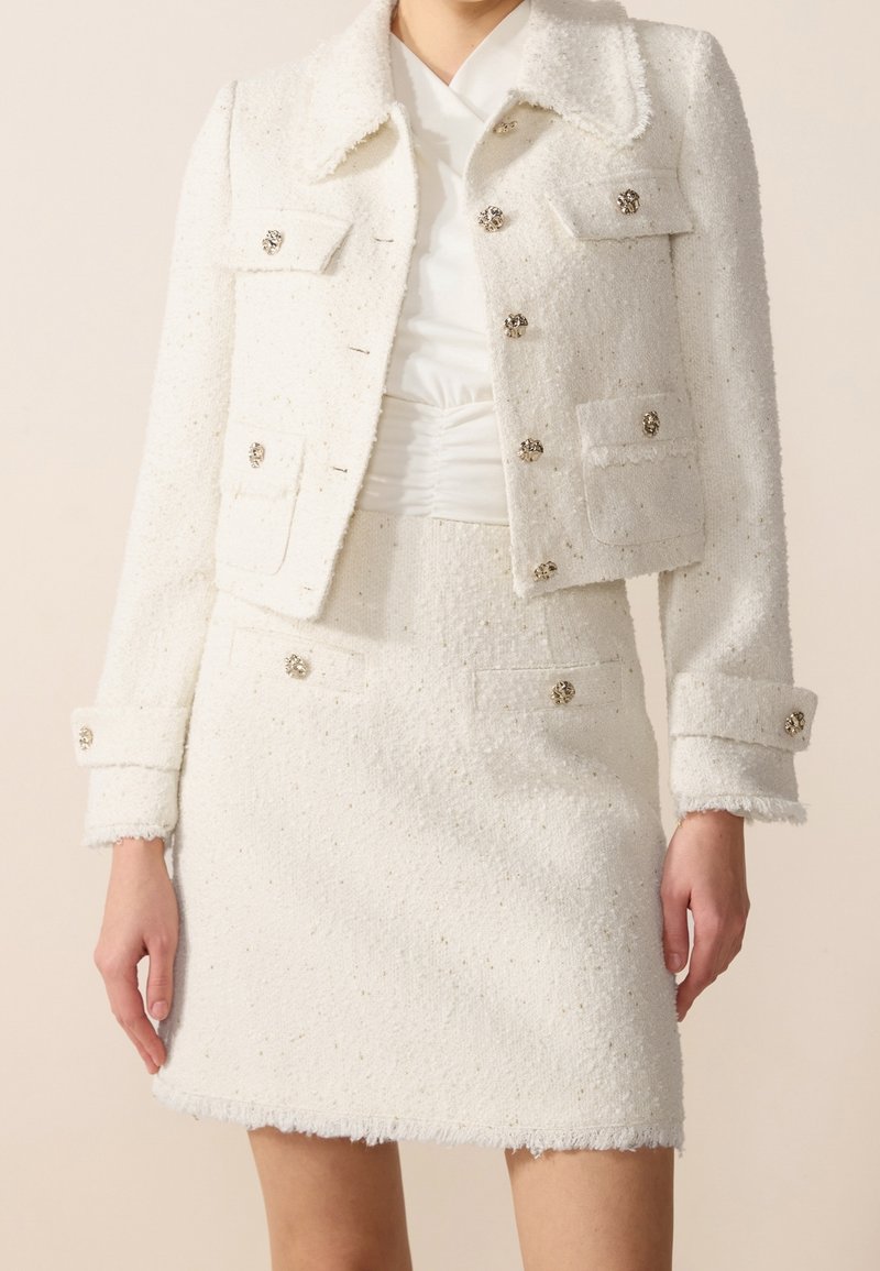 Femme portant un tailleur blanc texturé deux pièces avec des boutons dorés décoratifs et une jupe taille haute, debout devant un fond uni.