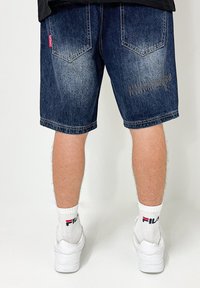 Denim shorts in dark blue with a distressed texture, featuring two back pockets and decorative stitching on the right leg. White socks and shoes visible.