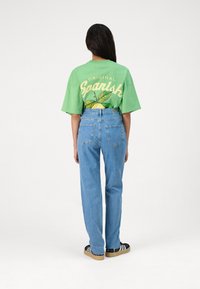 Camiseta verde de gran tamaño con un gráfico de "Original Spanish" y diseño de limón, combinada con jeans rectos de color azul claro y zapatillas negras.