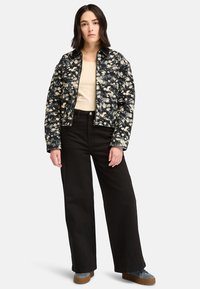 Chaqueta de mezclilla negra con estampado floral y cierre frontal, combinada con una blusa beige y pantalones de pierna ancha negros de cintura alta, y zapatillas de plataforma grises.