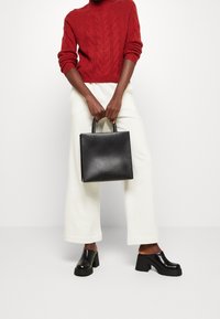 Personne portant un pull en laine tressé rouge, un pantalon blanc à jambes larges, des chaussures noires à talons épais et enfilables, tenant un sac à main en cuir noir.