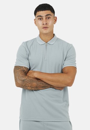 Młody mężczyzna o krótkich czarnych włosach i tatuażach na ramionach, ubrany w jasnoszarą koszulkę polo z zamkiem, ze skrzyżowanymi ramionami.