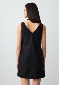 Schwarzes Leinenkleid mit V-förmigem Rücken, ärmellosem Design und subtiler Textur. Der Saum endet über dem Knie und weist ein zentrales Nahtdetail auf.