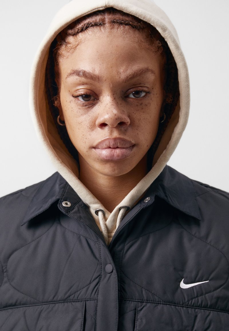 Junge Person mit Sommersprossen und geflochtenen Haaren, die einen beigen Hoodie unter einer schwarzen, gefütterten Nike-Jacke trägt und mit dem Gesicht zur Kamera vor einem weißen Hintergrund steht.