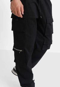 Pantalones cargo negros con múltiples bolsillos, detalles con cremallera y cordones ajustables en la cintura. Hechos de un tejido duradero.