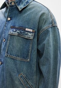 Jeans μπουφάν σε μπλε χρώμα με παραδοσιακό γιακά, διαθέτει μεγάλη μπροστινή τσέπη με ταμπελάκι λογότυπου, ορατά ραψίματα και άνετη εφαρμογή.