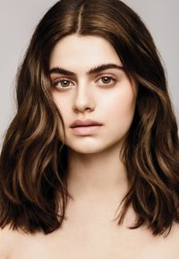 Retrato de una persona con cabello largo y ondulado de color marrón oscuro, piel clara y maquillaje natural. La iluminación suave resalta los rasgos faciales y la expresión.