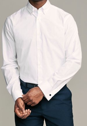 Homme portant une chemise blanche boutonnée avec des boutons de manchette et un pantalon bleu foncé ajustant son poignet gauche devant un fond gris uni.