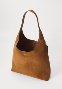 Bruine suède totebag met een brede opening, voorzien van een gladde leren hengsel en een aangehechte label, tegen een effen achtergrond.