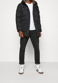 Sort polstret jakke med en hætte i kunstpels, lynlåslommer, brugt over en hvid t-shirt og sorte jeans, parret med sneakers.