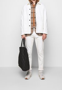 Chaqueta de lona blanca, camisa de cuadros marrón, pantalones blancos, zapatillas grises con acentos naranjas y un bolso de mano negro acolchado.