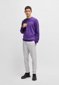 Lila sweatshirt med ribbade detaljer, grå byxor och svarta sneakers. Tröjan har en liten svart logotyp på bröstet.