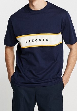 Marineblaues Baumwoll-T-Shirt mit einem weißen und gelben horizontalen Streifen, auf dem "LACOSTE" in Schwarz gedruckt ist. Kurze Ärmel und Rundhalsausschnitt.