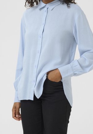 Henkilö, jolla on yllään vaaleansininen pitkähihainen kauluksellinen nappipaita, mustat housut ja toinen käsi taskussa, yksinkertaisen taustan edessä.