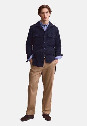 Giovane uomo che indossa una giacca blu navy sopra una camicia blu, pantaloni beige, scarpe scure e un maglione scuro legato in vita, in piedi con le mani nelle tasche.