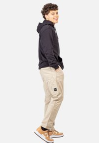 Junger Mann steht seitlich, trägt einen schwarzen Hoodie, beigefarbene Cargo-Hosen und tanfarbene sowie weiße Sneaker, lächelt mit den Händen in den Taschen.