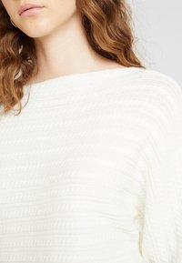 Pull en tricot blanc avec un motif texturé, présentant des rayures horizontales et un large col bateau, mettant en valeur une apparence douce et confortable.