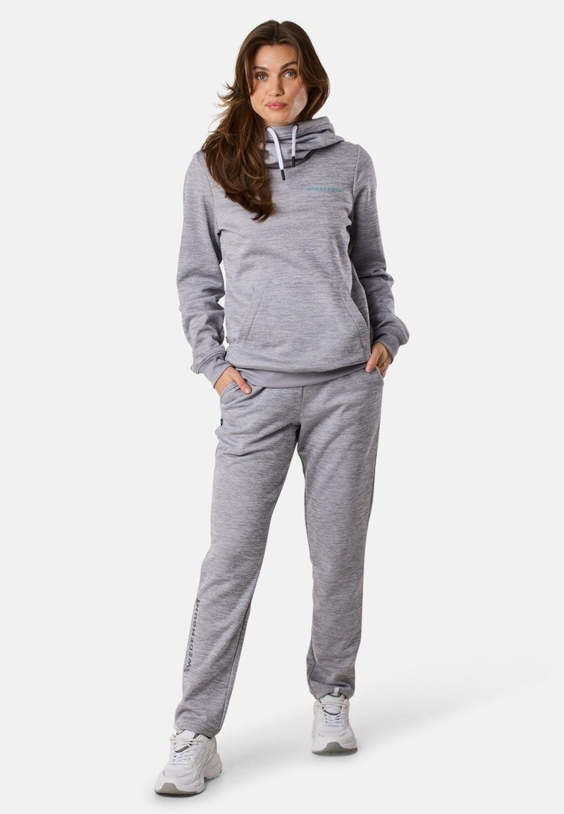 Kobieta stoi z rękami w kieszeniach, ubrana w dopasowany jasnoszary hoodie i dresy, z białymi sneakersami na białym tle.