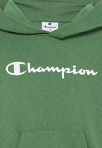 Grønn bomull hettegenser med lomme foran, som har en hvit "Champion"-logo i kursiv på brystet.