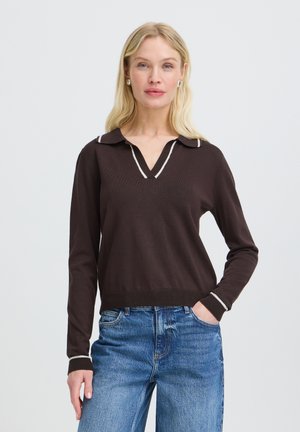 Femme blonde portant un pull à manches longues marron foncé avec une bordure blanche et un jean bleu, debout avec une main dans la poche devant un fond uni.