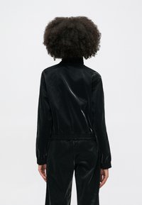 Juodas, blizgus švarkas su stovinčiu apykakle, elastingu liemeniu ir rankogaliais. Lygus paviršius su moderniu dizainu, atskleidžiančiu šiuolaikišką siluetą.