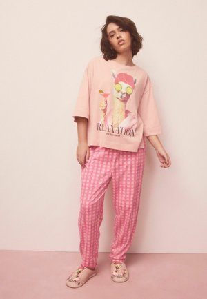 Femeie purtând pijama roz cu grafică de llama, ținând un cocktail, asortată cu pantaloni și papuci în carouri, pe fundal simplu.