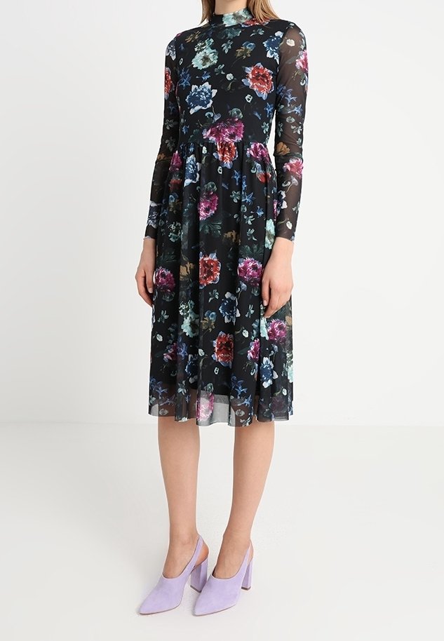 Robe noire à manches longues avec un motif floral en rouge, bleu et vert, dotée d'un corsage ajusté et d'une jupe évasée. Associée à des mules lilas.