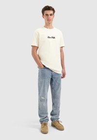 Jonge man die staat met zijn handen in zijn zakken, in een crèmekleurig T-shirt, lichte blauwe ripped jeans en tan veterschoenen.