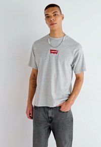 Grå kortærmet t-shirt med et rødt Levi's-logo på brystet, parret med mørkegrå jeans. Stoffet virker blødt og har en afslappet pasform.