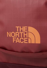 Piros textúrájú szövet, rácsos mintázattal, az "THE NORTH FACE" logó narancssárga színben, jól láthatóan a bal alsó sarokban elhelyezve.