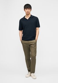 Νέος άνδρας που στέκεται με τα χέρια στις τσέπες, φοράει ένα υφασμένο μαύρο πόλο, ελαιδί παντελόνι και λευκά αθλητικά παπούτσια σε λευκό φόντο.