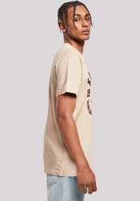 Beige cotton t-shirt with short sleeves, featuring a graphic print in navy and orange. Model stands sideways against a neutral background.
