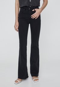 Jeans acampanados negros de mezclilla, con un diseño de cintura alta, cinco bolsillos y un acabado sin costuras. Combinados con sandalias blancas de punta abierta.