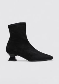 Bottine noire en suede avec un bout pointu et un petit talon angulaire. Présente un empiècement supérieur sans coutures semblable à une chaussette et une texture lisse.
