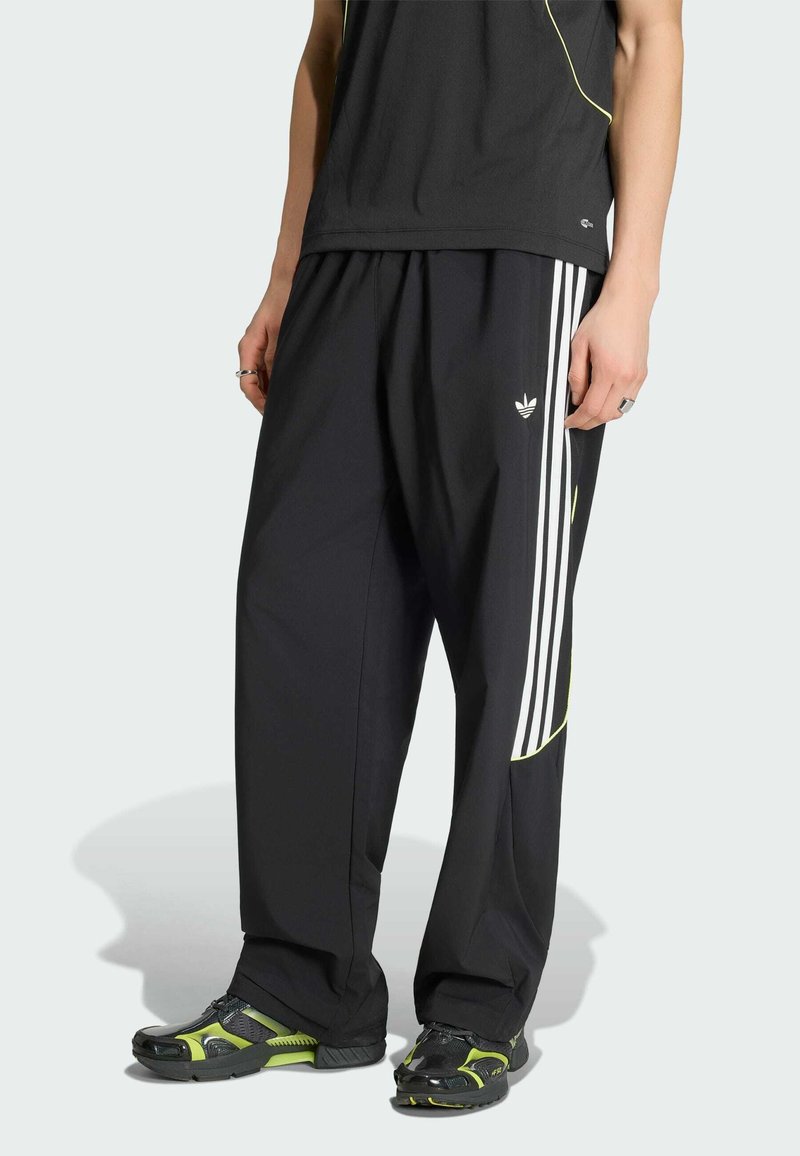 Personne portant un pantalon de survêtement Adidas noir avec des rayures blanches et des baskets noires avec des touches vertes, debout devant un fond clair uni.