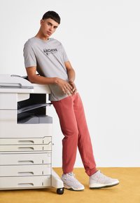 Maglietta grigia con grafica "ARCHIVE", abbinata a pantaloni tuta rossi e sneakers bianche, appoggiata a una stampante multifunzione.