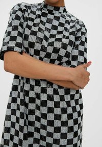 Schwarz-weiß kariertes Kleid mit Pailletten, hohem Kragen, kurzen Ärmeln und lockerem Schnitt. Auffällige glänzende Textur und geometrisches Muster.
