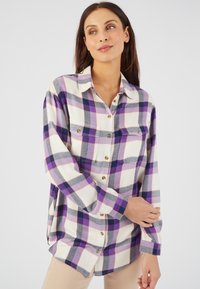 Camisa de franela en blanco con un patrón de cuadros en púrpura y gris. Presenta un cuello, cierre de botones y dos bolsillos en el pecho. Corte holgado con mangas largas.